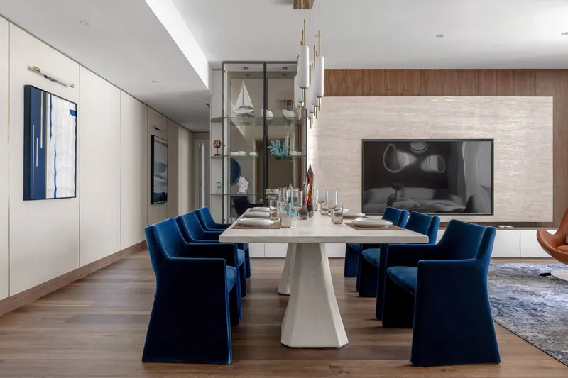 Dining area with a stone table, blue velvet chairs, a glass display cabinet and a travertine TV wall
